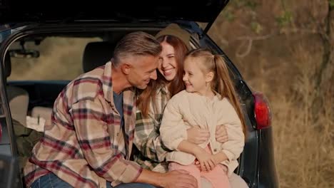 A-happy-middle-aged-brunette-man-with-gray-hair-in-a-checkered-brown-shirt-hugs-his-wife-and-little-daughter-in-the-open-trunk-of-a-black-car-during-his-summer-picnic-outside-the-city-in-the-evening