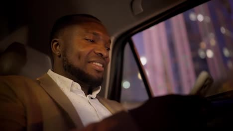 A-happy-Black-skinned-businessman-in-a-brown-suit-works-on-a-white-smartphone-and-watches-and-enjoys-the-views-of-the-evening-city-during-his-business-trip