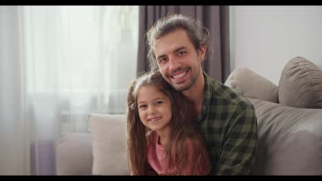 Portrait-of-a-Happy-father-of-a-brunette-man-in-a-Green-checkered-shirt-who-is-sitting-with-his-little-daughter,-a-brunette-girl-in-a-pink-dress-on-a-gray-sofa-and-hugging-in-a-modern-apartment