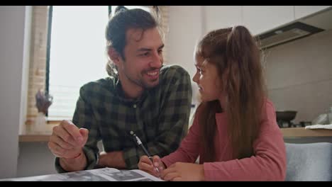 Happy-brunette-man-in-a-green-checkered-shirt-with-his-little-daughter-in-a-pink-dress-doing-homework-and-teaching-his-daughter-new-things-while-sitting-in-the-kitchen-at-the-table-in-a-modern-apartment