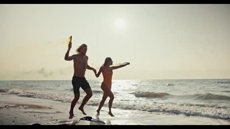 A-blond-guy-holds-his-blonde-girlfriend-in-an-orange-swimsuit-and-they-run-along-the-orange-brown-seashore-holding-smoky-fireworks-in-their-hands-that-leave-behind-green-and-yellow-colors-on-the-seashore-in-the-morning