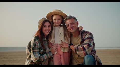 Portrait-of-a-happy-family,-a-little-blonde-girl-in-a-Straw-hat-stands-in-the-middle-and-is-hugged-from-different-sides.-Her-father-is-a-brunette-man-with-gray-hair-in-a-checkered-shirt-and-her-mother-is-a-girl-and-a-brunette-in-a-Green-checkered-shirt-on-a-deserted-shore-during-a-summer-hike-outsid