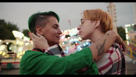 Close-up-shot-of-a-blonde-girl-with-short-hair-with-glasses-in-a-plaid-shirt-and-a-girl-with-short-green-hair-in-a-Green-shirt-hug-each-other-and-stroke-their-shoulders-during-their-date-in-the-amusement-park