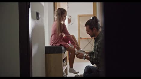 Shot-from-the-side-of-a-brunette-man-in-a-checkered-green-shirt.-A-lonely-father-helps-his-little-daughter-in-a-pink-dress-put-on-white-shoes-before-starting-their-walk.-A-brunette-man-helps-his-daughter-put-on-white-shoes-in-the-hallway-of-a-modern-apartment