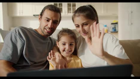 Happy-family,-a-brunette-guy-with-stubble,-his-brunette-girlfriend-in-a-white-T-shirt-and-their-joyful-daughter-in-a-yellow-dress-communicate-with-their-family-and-friends-via-video-conference-using-a-laptop-in-a-modern-studio-room