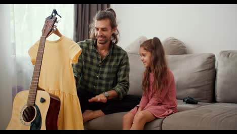 A-little-brunette-girl-in-a-pink-dress-sits-on-the-sofa-near-her-father-who-steams-a-pink-dress-using-a-special-steamer-in-a-modern-apartment.-Single-father-brunette-man-in-a-green-checkered-shirt-steams-his-daughter's-yellow-dress-at-home