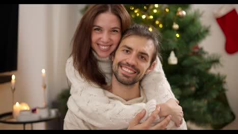 Portrait-of-a-Happy-guy-and-a-brunette-girl-in-a-White-sweater-who-hugs-her-boyfriend-from-behind-and-smiles-in-a-cozy-Christmas-decorated-room-with-a-Christmas-tree