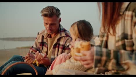 A-middle-aged-brunette-man-in-a-checkered-shirt-peels-a-banana-fruit-for-his-little-daughter-during-a-picnic-outside-the-city-in-summer.-Happy-man-feeding-his-little-daughter-with-his-wife-during-a-picnic-outside-the-city-in-summer