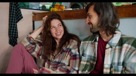 Happy-brunette-girl-in-a-plaid-shirt-sits-on-the-floor-in-her-trailer-and-communicates-with-her-brunette-boyfriend-with-stubble-in-a-green-plaid-shirt-and-a-white-T-shirt-during-her-vacation-outside-the-city-near-a-motor-home-in-a-camp-in-the-summer