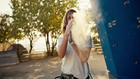 Portrait-of-a-happy-blonde-girl-climber-who-takes-sports-chalk-out-of-her-bag-and-rubs-it-in-her-hands-near-a-blue-climbing-wall.-White-sports-chalk-scatters-in-a-light-haze-along-the-hands-of-a-girl-climber-on-a-sunny-summer-day