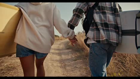 A-blond-guy-in-a-checkered-shirt-walks-hand-in-hand-with-his-blonde-girlfriend-in-a-white-sweatshirt-and-they-hold-surfboards-in-their-hands-and-walk-along-a-path-near-dried-grass-and-green-trees