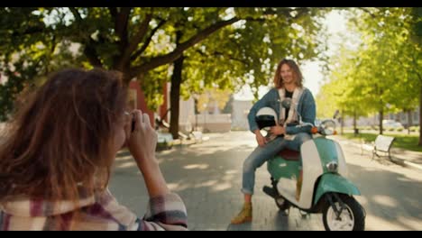 Over-her-shoulder,-a-brunette-girl-in-a-checkered-shirt-uses-a-camera-to-photograph-her-boyfriend-with-long-curly-hair-on-a-green-moped-and-shows-him-how-to-pose-correctly-in-a-city-summer-park