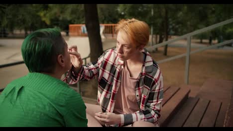 Over-the-shoulder:-A-girl-with-short-dark-yellow-hair-in-a-pink-checkered-shirt-puts-glitter-on-the-face-of-her-partner-a-girl-with-short-hair-with-green-hair-in-a-green-shirt-on-brown-stands-benches-near-a-skate-park-in-a-green-park-in-summer