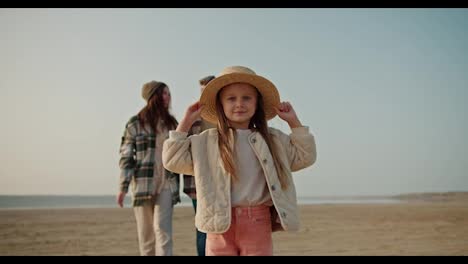 portrait-of-a-happy-little-blonde-girl-in-a-Straw-hat-who-looks-at-the-camera-and-smiles-and-her-parents-come-up-to-her-from-behind-and-hug-her,-posing-with-her-on-a-deserted-seashore-during-a-picnic-on-a-hike