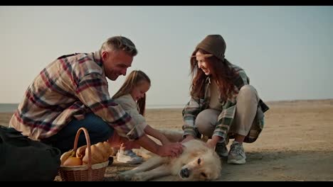 Happy-family-with-their-pet-dog-on-a-picnic.-Happy-brunette-man-in-a-brown-checkered-shirt-with-his-wife-and-daughter-petting-a-large-cream-colored-dog-during-their-picnic-on-a-deserted-seashore-in-summer