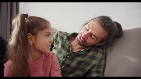 A-brunette-man-with-stubble-in-a-checkered-green-shirt-sits-on-the-sofa-and-communicates-with-his-little-brunette-daughter-in-a-pink-jacket-in-a-modern-home.-Happy-brunette-man-single-father-communicates-with-his-little-daughter-while-sitting-on-a-gray-sofa-in-a-modern-apartment