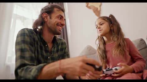 Close-up-shot-of-a-brunette-man-in-a-green-checkered-shirt-playing-and-telling-how-to-properly-hold-a-joystick-in-his-hands-together-with-his-daughter,-a-little-brunette-girl-in-a-pink-dress-on-a-gray-sofa-in-a-modern-apartment