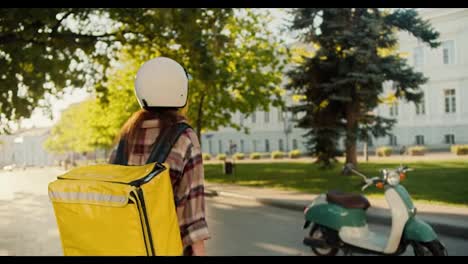 Vista-Trasera-De-Una-Chica-Mensajera-Con-Una-Camisa-A-Cuadros,-Un-Casco-Blanco-Y-Una-Gran-Bolsa-Amarilla,-Se-Dirige-A-Su-Ciclomotor,-Se-Sube-A-él-Y-Comienza-A-Moverse-Para-Llevar-El-Pedido-En-La-Calle-Cerca-Del-Parque-En-Un-Clima-Soleado-En-Verano.