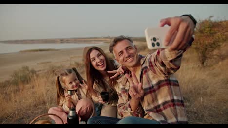 Un-Hombre-Moreno-Feliz-Con-Cabello-Gris-Y-Una-Camisa-A-Cuadros-Se-Toma-Una-Selfie-Usando-Un-Teléfono-Con-Su-Esposa-Y-Su-Pequeña-Hija-Durante-Su-Picnic-Fuera-De-La-Ciudad-En-Verano.