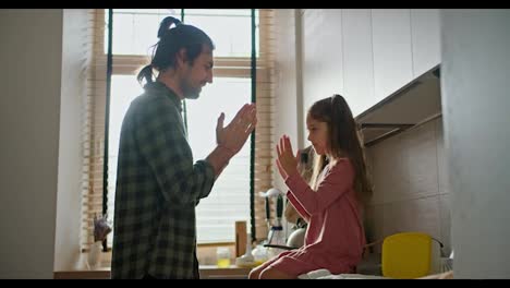 Happy-father,-a-brunette-man-in-a-checkered-green-shirt-plays-with-his-little-brunette-daughter-in-a-pink-dress,-clapping-his-hands-while-in-a-modern-kitchen