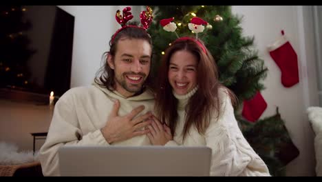 Happy-guy-and-girl-in-New-Year's-hoops-and-white-sweaters-sit-in-front-of-a-gray-laptop-and-congratulate-their-friends-and-family-Happy-New-Year-near-the-New-Year-tree-in-a-cozy-room-in-a-winter-evening