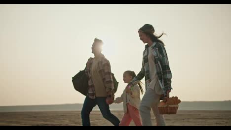 Side-view-of-a-happy-brunette-girl-in-a-green-checkered-shirt-walking-and-holding-hands-with-her-little-daughter-and-husband-during-her-hike-and-picnic-in-the-summer