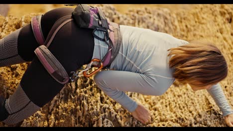 Vertical-video-of-a-blonde-girl-with-a-bob-hairstyle-in-a-gray-T-shirt-in-the-necessary-equipment-for-cleaving-and-using-a-safety-net,-climbs-up-a-yellow-rock