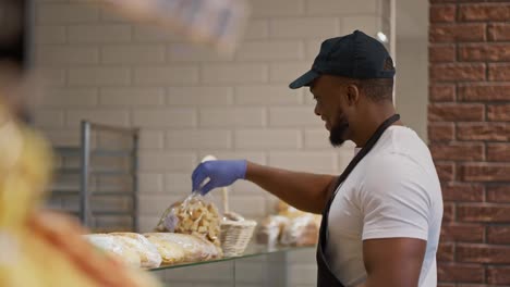 Side-view-of-a-Black-skinned-man-happy-supermarket-worker-in-a-white-t-shirt-and-black-cap-laying-out-pastries-and-other-bakery-products-on-a-glass-display-case-in-a-modern-grocery-supermarket