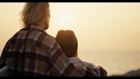 Rear-view-of-a-happy-blond-guy-in-a-plaid-shirt-and-his-blond-girlfriend-in-a-black-hat-and-plaid-shirt-standing-and-hugging-and-looking-at-the-sea-in-the-morning-at-Sunrise