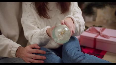 Close-up-shot-of-a-brunette-girl-in-a-white-sweater-shaking-a-glass-New-Year's-sphere-and-her-boyfriend-putting-his-hand-on-her-lap-during-their-gatherings-and-communication-in-a-cozy-New-Year's-decorated-room