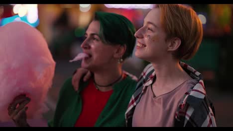 Happy-blonde-girl-with-glitter-on-her-face-in-a-checkered-pink-t-shirt-and-a-girl-with-short-green-hair-eating-a-large-pink-cotton-candy-and-looking-at-the-bright-rides-in-the-amusement-park-during-their-LGBT-date-in-the-evening