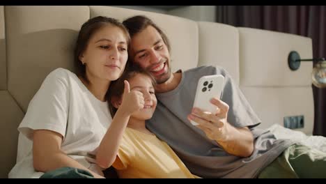 A-brunette-man-in-a-gray-T-shirt-takes-a-selfie-with-his-wife-and-little-brunette-daughter-in-a-yellow-dress-using-a-white-phone-on-a-light-brown-bed-in-a-modern-apartment