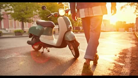 Close-up-shot-of-a-guy-in-denim-walking-up-to-his-green-moped,-getting-on-it-and-driving-off-on-the-street-during-sunrise-in-summer.-The-man-with-curly-hair-in-a-helmet-sits-on-a-moped-during-sunny-weather-in-summer