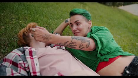 Close-up-shot-of-a-girl-with-a-short-haircut-with-green-hair-in-a-checkered-green-shirt-stroking-her-girlfriend-a-blonde-with-a-short-haircut-wearing-glasses-in-a-checkered-pink-shirt-during-her-date-in-the-park-lying-on-the-lawn-on-green-grass-in-the-summer