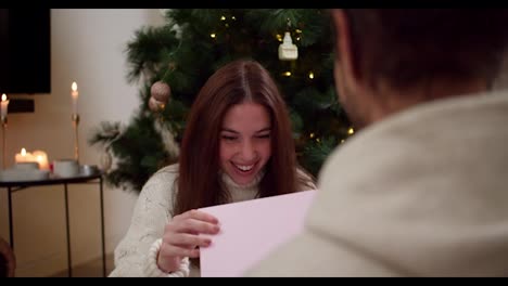 Over-the-shoulder-the-brunette-girl-opens-the-pink-box-and-looks-at-the-gift-that-her-boyfriend-gave-her-and-hugs-him-in-the-Christmas-atmosphere-at-home