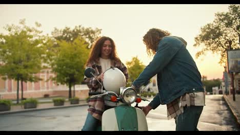 The-girl-turns-on-and-shirts-with-curly-brown-hair-gets-on-her-green-moped,-puts-on-a-white-helmet,-her-boyfriend-with-curly-hair-in-a-denim-shirt-walks-next-to-her,-puts-an-olive-helmet-on-his-head-and-does-not-start-riding-a-moped-on-the-street