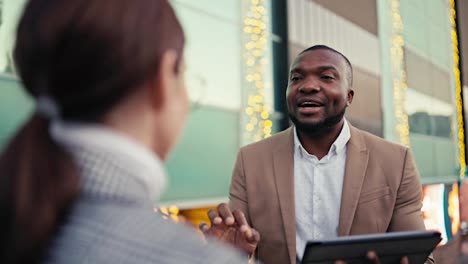 Over-the-shoulder-a-man-with-Black-skin-with-a-short-haircut-and-a-beard-in-a-brown-suit-a-businessman-talks-to-a-brunette-girl-in-a-gray-jacket-and-holds-a-large-black-tablet-in-his-left-hand-in-the-city-near-a-building