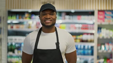 Retrato-De-Un-Hombre-Feliz-De-Piel-Negra-Con-Una-Camiseta-Blanca-Y-Un-Delantal-Negro-Que-Sonríe-Y-Posa-Cerca-De-Los-Mostradores-De-Comida-En-Un-Supermercado-Moderno.