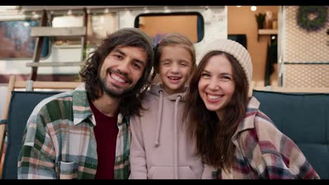 Retrato-De-Una-Familia-Feliz,-Un-Chico-Moreno-Con-Barba-Incipiente-Con-Una-Camisa-A-Cuadros-Verde,-Una-Mujer-Morena-Con-Una-Camisa-A-Cuadros-Y-Su-Pequeña-Hija,-Una-Niña-Rubia-Con-Una-Sudadera-Con-Capucha-Rosa,-De-Pie-Cerca-De-Su-Caravana,-Sonriendo-Y-Mirando-A-La-Cámara-Durante-Su-Picnic-Fuera-De-La-Ciudad-En-Verano.