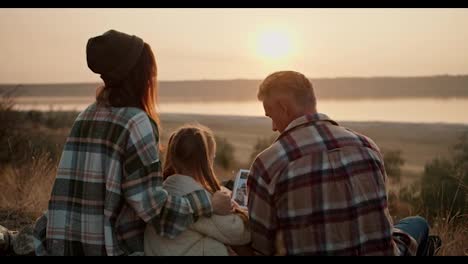 Close-up-shot-of-a-happy-brunette-girl-in-a-green-checkered-shirt-sitting-on-a-mat-with-her-middle-aged-man-husband-and-little-daughter,-they-look-at-the-sunset-and-enjoy-the-view-from-the-hill-during-their-picnic-outside-the-city