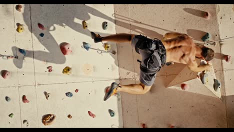 Vertical-video-of-a-brunette-guy-in-Gray-shorts-with-a-bare,-sculpted-torso-climbs-up-the-route-of-a-climbing-wall-and-hangs-on-one-of-the-ledges-with-the-help-of-a-belay-and-his-strong-arms-during-a-rock-climbing-training-at-a-climbing-wall-in-the-summer