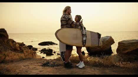 Romantic-meeting-of-a-blond-guy-with-a-beard-and-his-blonde-girlfriend-in-a-checkered-shirt,-who-holds-a-surfboard-and-hugs-her-boyfriend-on-a-rocky-shore-near-the-sea