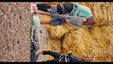 Vertical-video-of-a-girl-with-a-bob-hairstyle,-a-brunette-guy-in-a-gray-T-shirt-and-a-blonde-girl-in-a-black-sports-summer-uniform-attaching-special-equipment-and-insurance-to-their-belt-for-rock-climbing-against-the-backdrop-of-yellow-rocks-that-they-are-going-to-climb