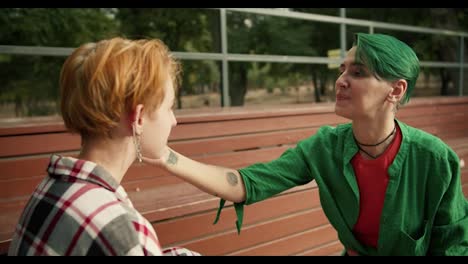 A-girl-with-short-green-hair-in-a-green-shirt-makes-makeup-for-her-friend-with-short-orange-hair-in-a-checkered-shirt-on-brown-benches-in-the-park.-Lesbian-couple-of-LGBT-girls-in-bright-clothes-and-bright-appearance-in-the-park-doing-make-up