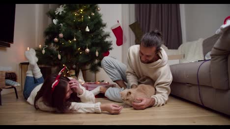 A-brunette-guy-in-a-white-sweater-together-with-his-brunette-girlfriend-in-a-white-sweater-are-sitting-on-the-floor-and-playing-with-their-cream-colored-cat-near-a-decorated-New-Year's-tree-in-a-cozy-house-with-a-New-Year's-atmosphere-in-winter