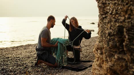 A-blonde-girl-with-curly-hair-in-black-clothes-untangles-and-unties-a-special-rope-together-with-her-boyfriend-in-order-to-start-rock-climbing-on-a-rocky-shore-near-the-rocks