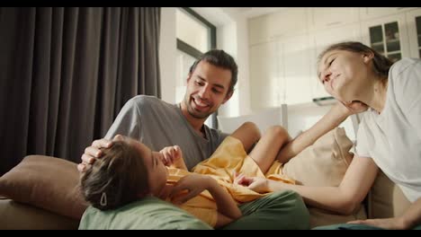 A-little-brunette-girl-in-a-yellow-dress-lies-on-her-dad-s-lap-and-communicates-with-her-parents-while-sitting-on-a-light-brown-in-a-modern-room