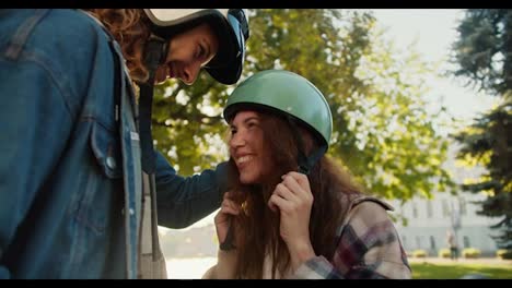 Close-up-shot-of-a-happy-brunette-girl-in-a-green-moped-helmet-in-a-plaid-shirt-hugging-and-looking-at-her-boyfriend-with-curly-hair-in-a-denim-jacket-in-a-city-summer-park