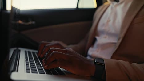 Close-up-shot-of-a-man-Businessman-with-Black-skin-color-and-in-a-brown-jacket-works-and-types-on-a-laptop-while-driving-in-the-passenger-seat-in-a-modern-car-interior