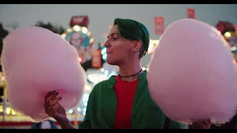 A-lesbian-girl-with-a-short-green-haircut-in-a-Green-shirt-holds-huge-pink-cotton-candy-in-her-hands-and-bites-into-it-during-her-walk-in-a-bright-amusement-park